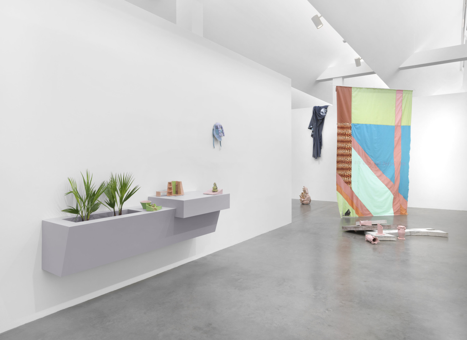 Hinten rechts sieht man ein aufgespanntes Stück Stoff, das übermenschgroß ist. Darauf mehrere bunte Felde, die durch lachsfarbene dickere Streifen unterbrochen werden. Davor liegen rosafarbene Gefäße, die teilweise zerbrochen sind