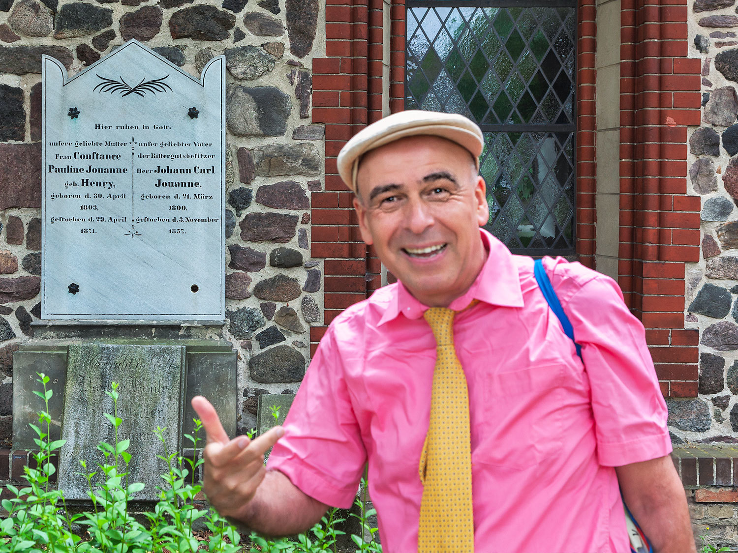 Auf der rechten Seite ein Mann in einem pinken Hemd mit einer gelben Krawatte. Er hat eine Schiebermütze auf. Dahinter eine gebauerte Wand aus Feldsteinen