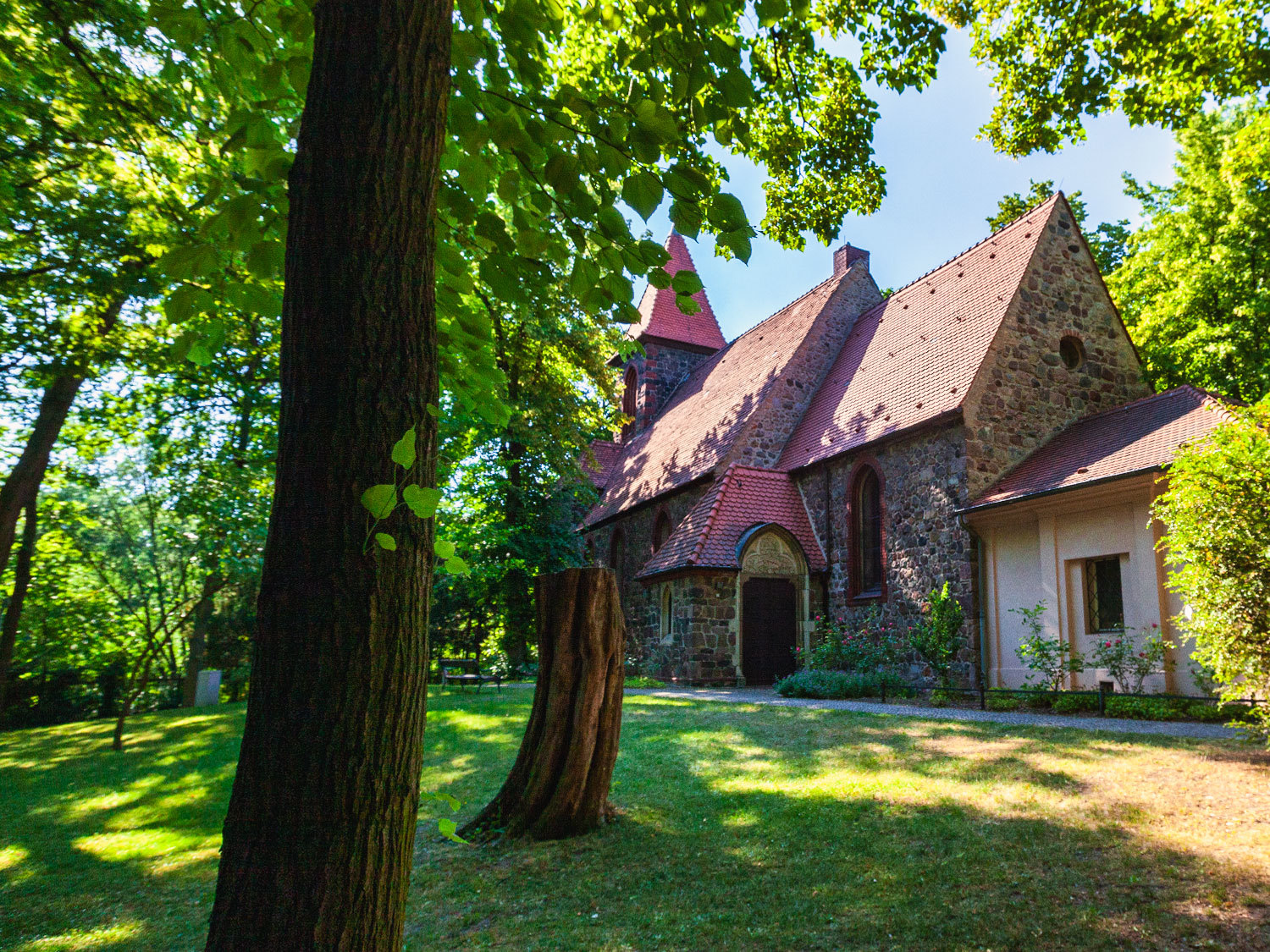 Auf der rechten Bildseite eine aus Feldsteinen gemauerte Kirche und eine roten Dach. Die Kirche steht in einem Park mit Laubbäumen.