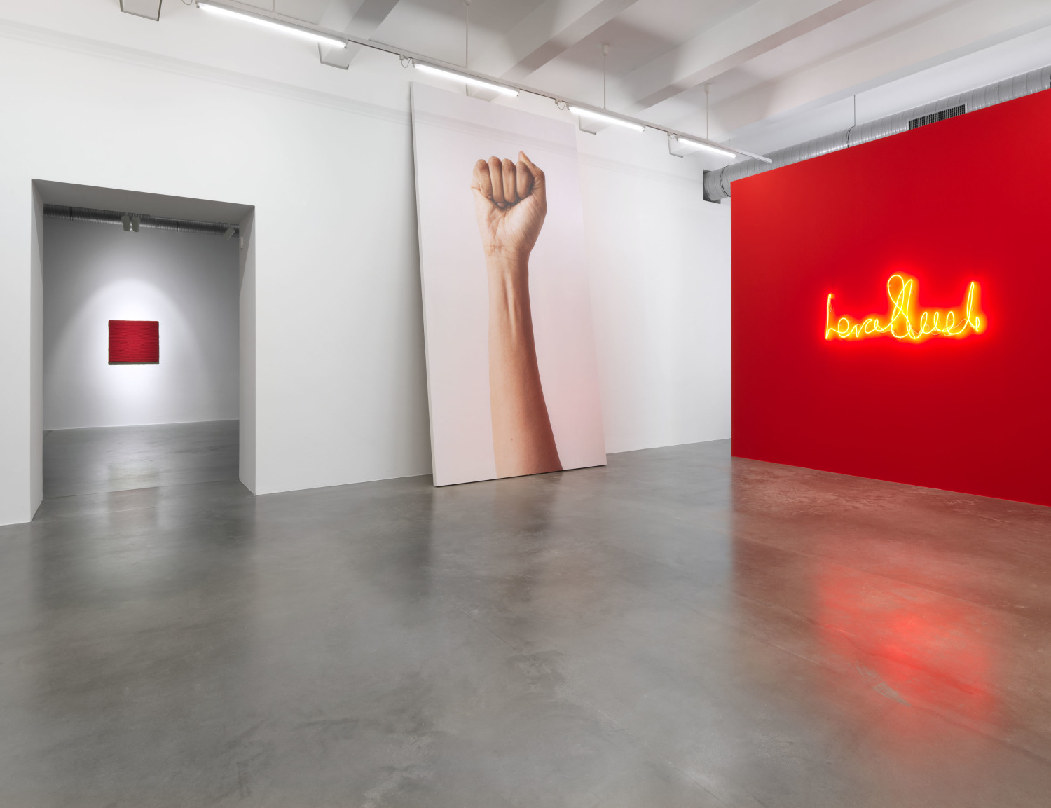 In einem weißen Raum mit einem Betonfußboden sieht man auf der rechten Seite eine rote Wand mit einem leuchtenden Schriftzug. Links daneben ein übermenschgroßes Bild eines ausgestreckten Arms mit einer geballten Faust