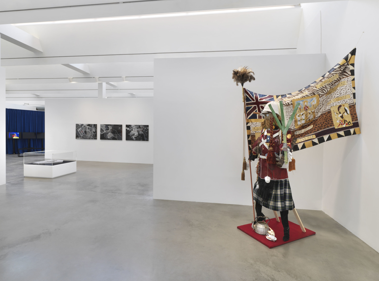 Auf der rechten Bildseite eine Schaufensterpuppe, die einen Schottenrock trägt, eine rote Jacke mit weißen Schnüren darüber, die an eine Millitäruniform erinnern soll. Die Puppe hat einen roten Bart und trägt einen weißen Helm. Hinter ihr eine Fahne mit einer kleineren britischen Fahne darauf und vielen afrikanischen Symbolen und Mustern, wie diversen Tierprinten