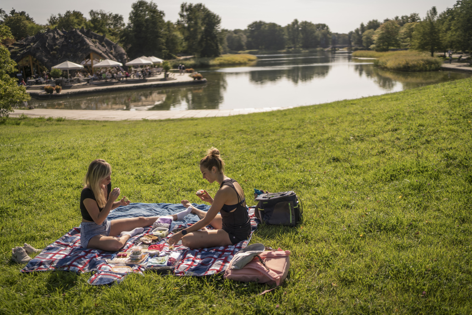Eine grüne Wiese. Auf einer Picknick-Decke sitzen zwei Frauen sich gegenüber und unterhalten sich. Sie haben Kleinigkeiten zum Essen dabei. Dahinter ein See