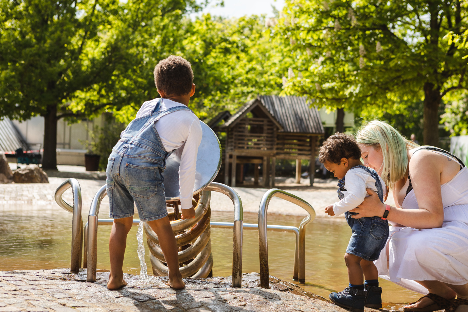 Zwei kleine Jungs spielen am Wasserspielplatz. Eine Frau mit blonden Haare hält den kleineren der beiden Jungen gerade fest. Im Hintergrund grünbelaubte Bäume