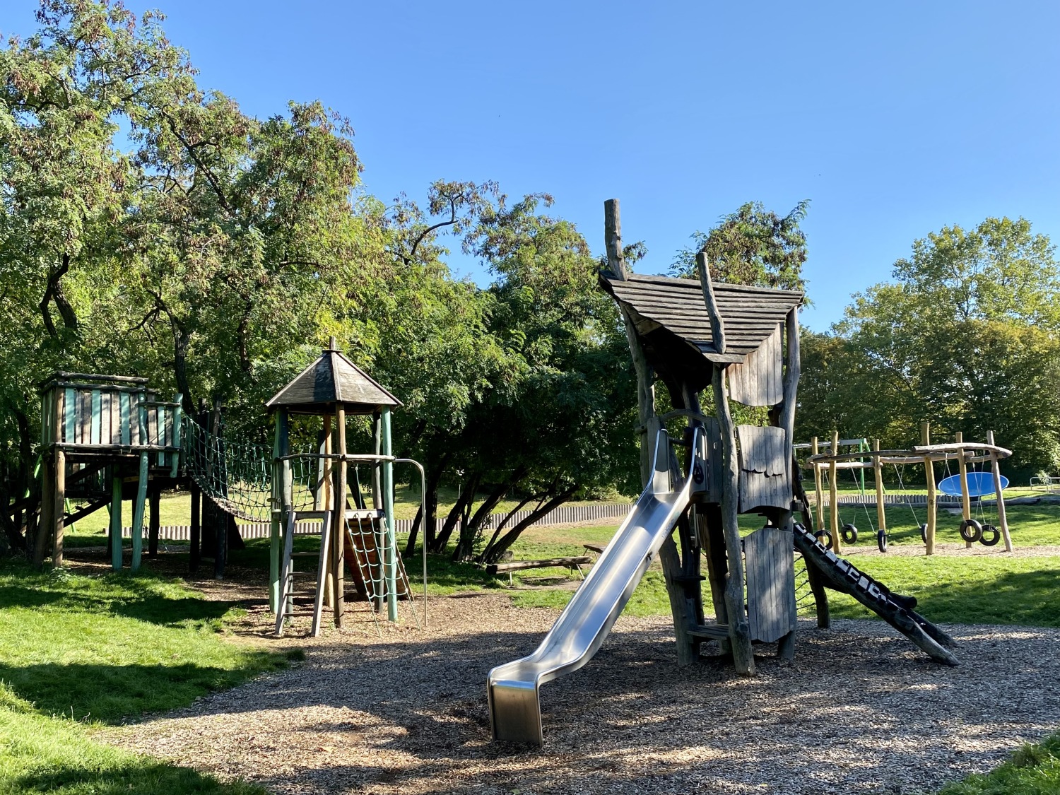 Auf der rechten Bildseite ein Turm, der aus Holz gebaut wurde. Vom Podest führt eine silberfarbene Rutsche nach unten. Rechts hinter dem Turm ein Gerüst, aus Holzstämmen, an dem sechs Reifenschaukeln befestigt sind. Dahinter eine blaue Drehscheibe. Auf der linken Bildseite ein weiterer Holzturm. Dieser ist über eine Hängebrücke mit einer weiteren hölzernen Plattform verbunden. Überall auf dem Spielplatz verteilt stehen Bäume mit grünem Laub. Untern den Spielgeräten liegt jeweils dunkelbrauner Rindenmulch.