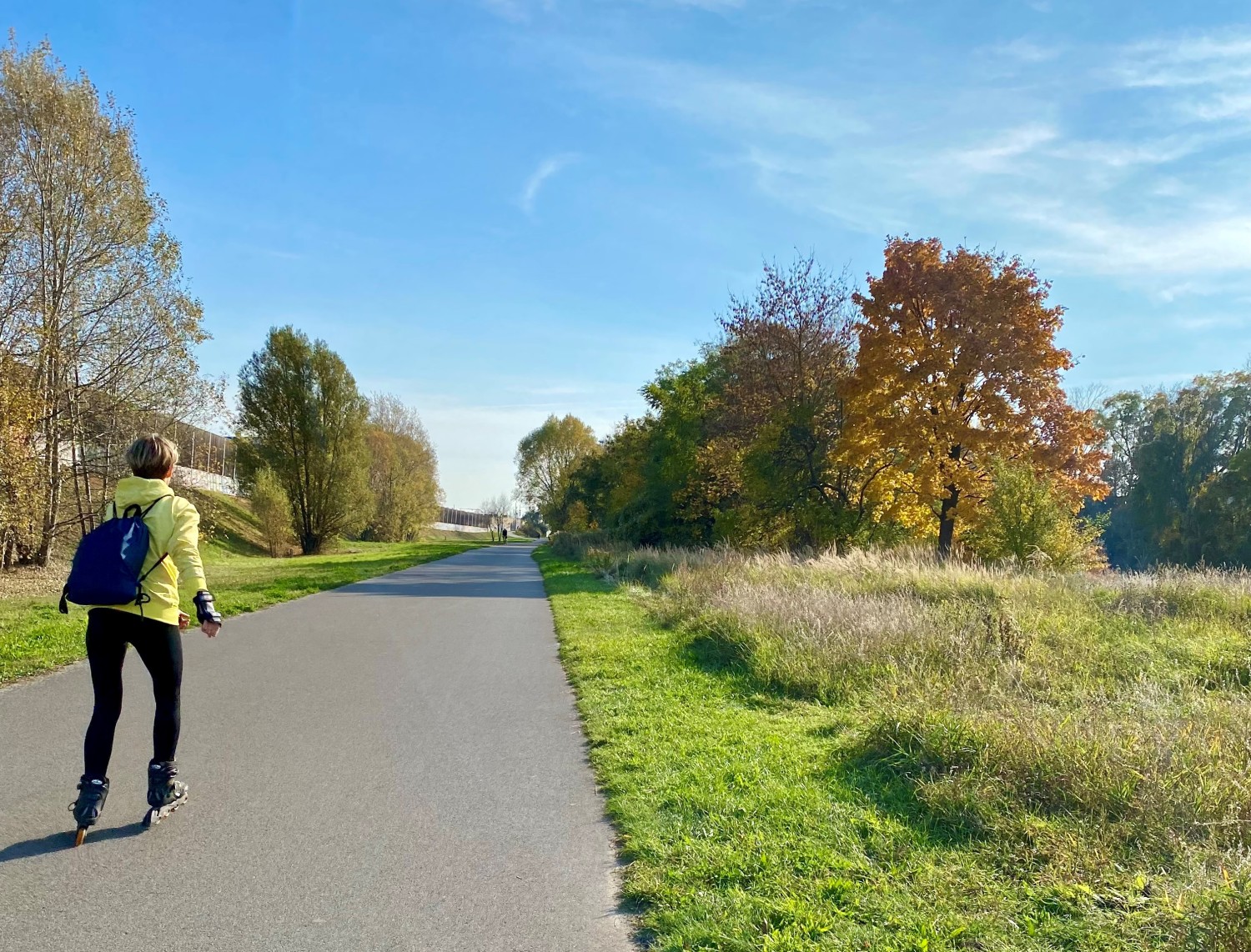 Ein asphaltierter Weg in der Bildmitte. Eine Frau ist auf diesem Weg mit Inlinern unterwegs. Man sihet sie von hinten. Sie hat eine gelbe Jacke an und eine schwarze Hose. Sie hat einen schwarzen Turnbeutel auf dem Rücken und hat kurze blonde Haare. Rechts und links des Weges ist Wiese. Auf der Wiese wachsen Bäume, deren Blätter schon in rot, gelb und braun herbstlich eingefärbt sind. Der Himmel ist blau mit leichten Schleierwolken