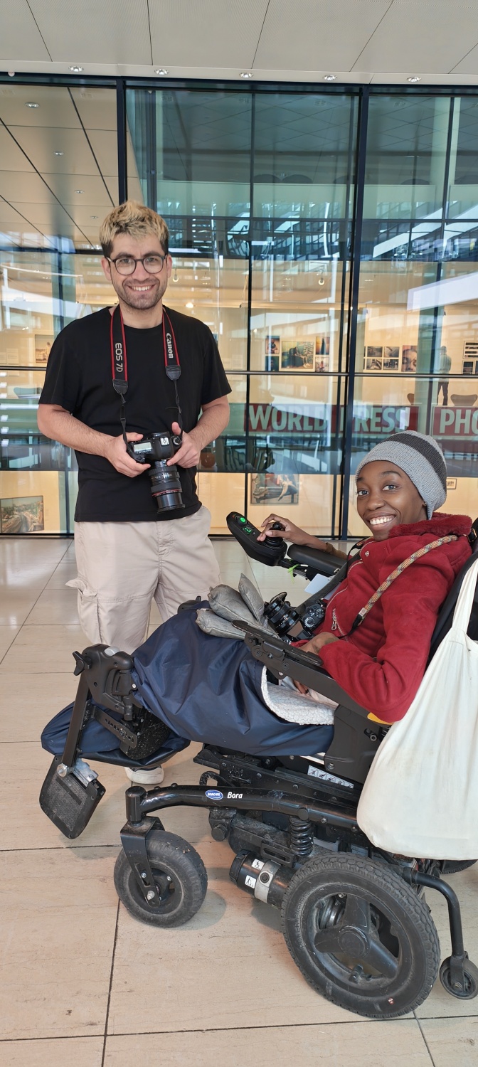 Eine stehende Person und eine Person, die einen Rollstuhl nutzt, befinden sich mit umgehängten Kameras in einer Galerie.
